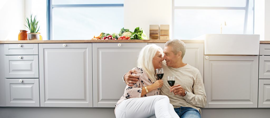 Two elderly people sitting on the kitchen floor with wine glasses, sharing an intimate moment